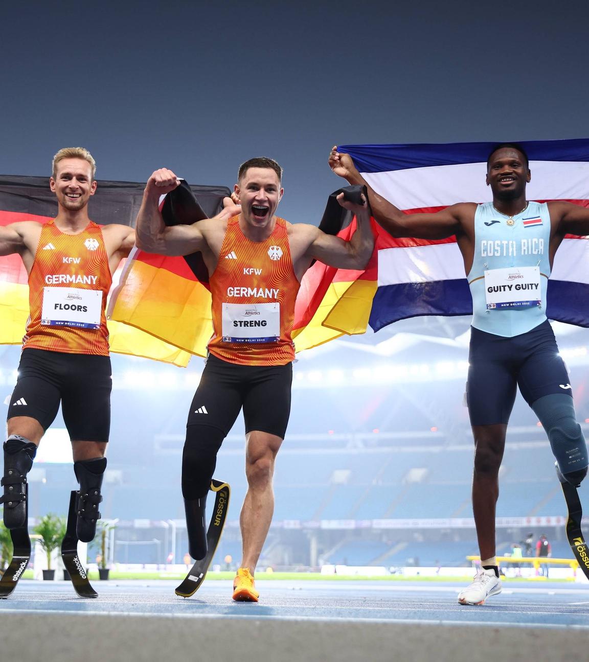 Johannes Floors und Felix Streng (Team Deutschland) sowie Sherman Guity Guity (Costa Rica) nach dem 100-Meter-Finale (T64) bei der Para-WM in Neu-Delhi am 30.09.2025.