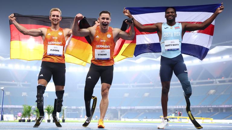 Johannes Floors und Felix Streng (Team Deutschland) sowie Sherman Guity Guity (Costa Rica) nach dem 100-Meter-Finale (T64) bei der Para-WM in Neu-Delhi am 30.09.2025.