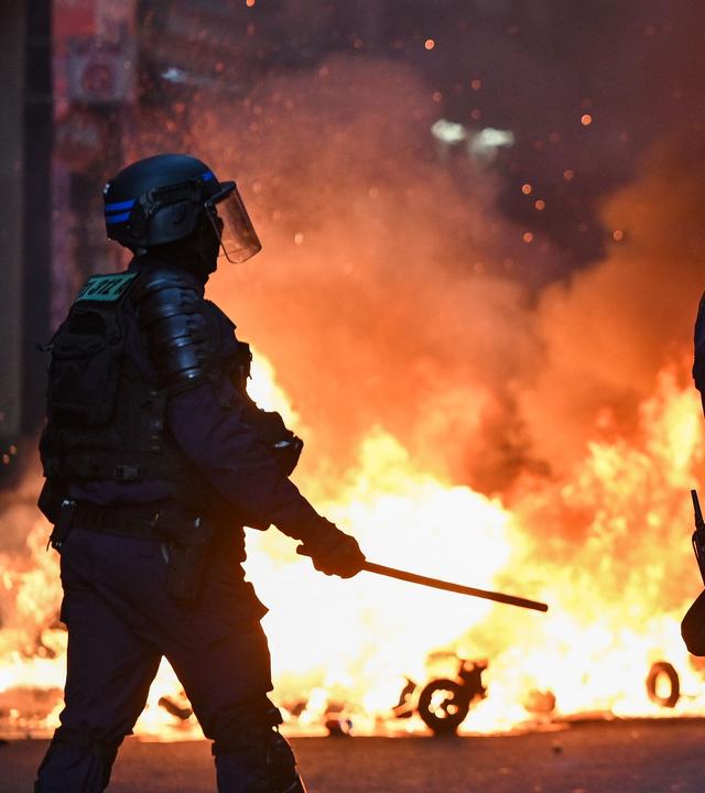 Zwei Polizisten stehen am Abend vor brennenden Gegenständen auf einer Straße. 