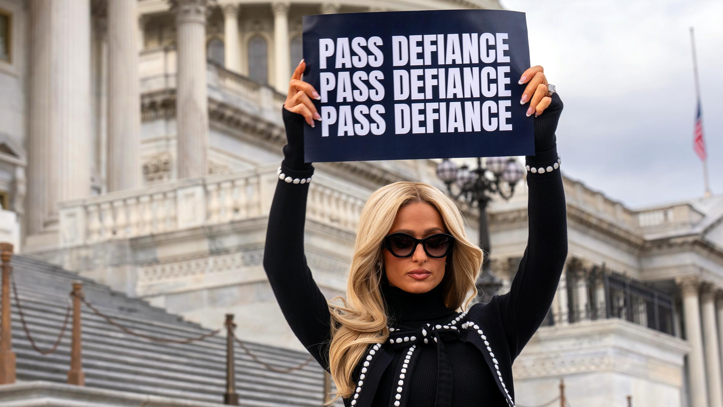 Paris Hilton hält ein Schild mit der Aufschrift "Pass Defiance".