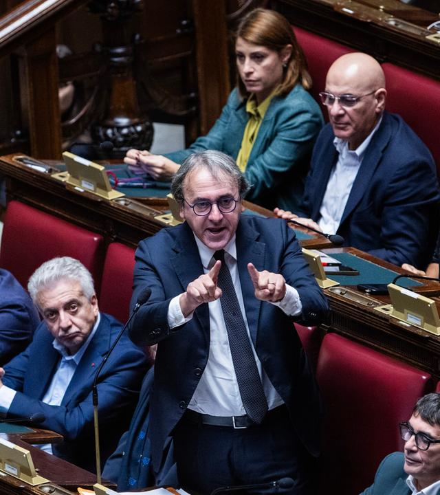 Mitglieder des italienisches Parlaments. Ein Mann steht mit erhobenen Zeigefingern an seinem Sitz.
