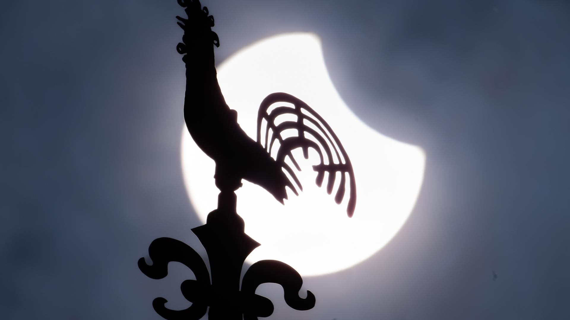 Die zum Teil durch den Mond verdeckte Sonne steht hinter dem Wetterhahn der Kreuzkirche in Frankfurt-Preungesheim am Himmel.