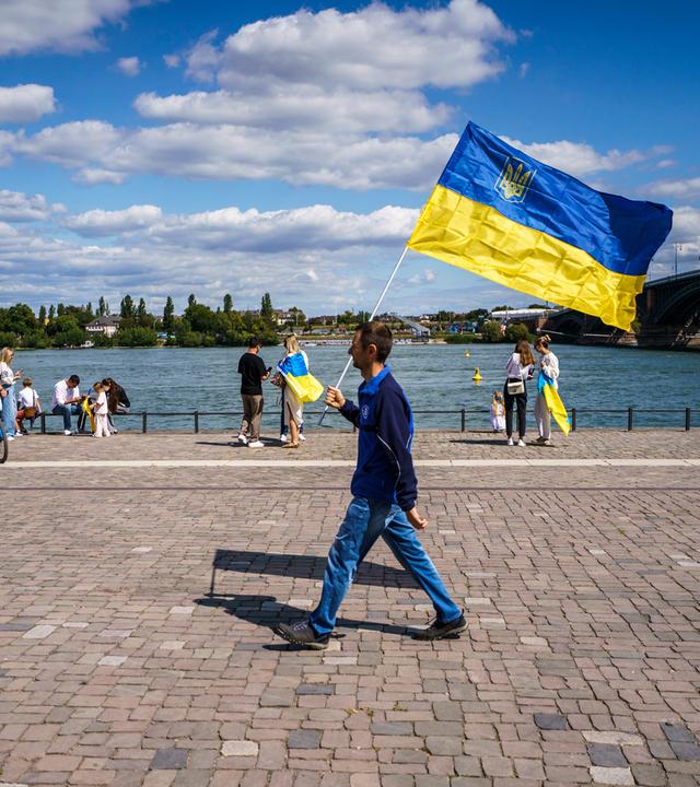 Ein Mann geht auf einer Kundgebung zum 34. Unabhängigkeitstag der Ukraine mit der ukrainischen Nationalflagge am Rheinufer.