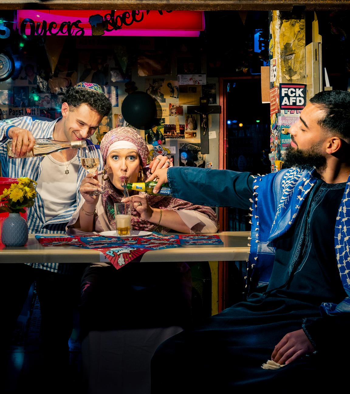 Atheistin Josi sitzt hinter der Bar und trinkt muslimisches Bier aus einem Glas. Vor ihr sitzt Muslim Omar, der ihr das muslimische Bier einschenkt. In der anderen Hand hält Josi ein Weinglas. Dieses füllt ihr Jude Lars auf.