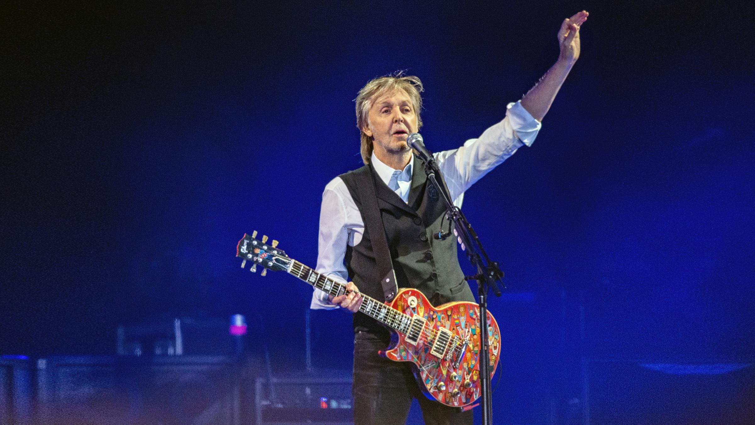 Paul McCartney steht mit einer Gitarre auf der Bühne und spricht ins Mikrofon.