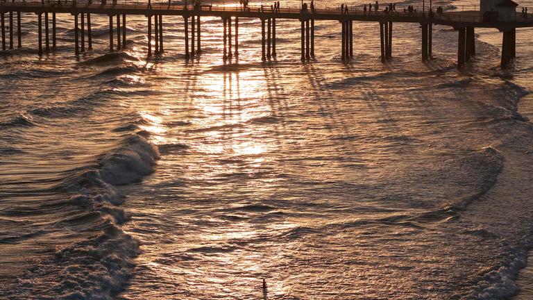 Symbolbild: Eine Luftaufnahme zeigt Menschen auf dem Pier von Huntington Beach über dem Pazifik, während eine Person im Wasser watet (Aufnahme vom 1. Juli 2026 in Huntington Beach, Kalifornien).