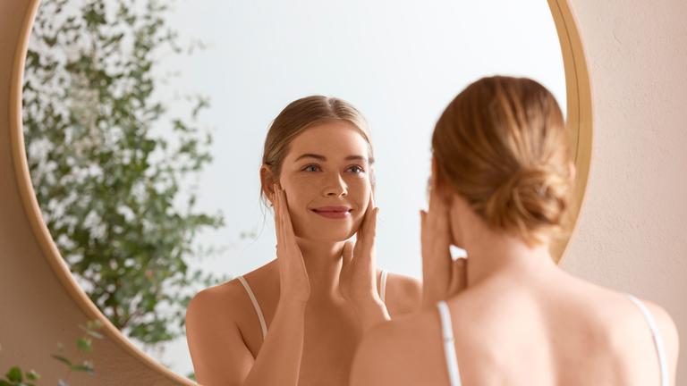 Eine junge Frau begutachtet ihr Gesicht im Spiegel.