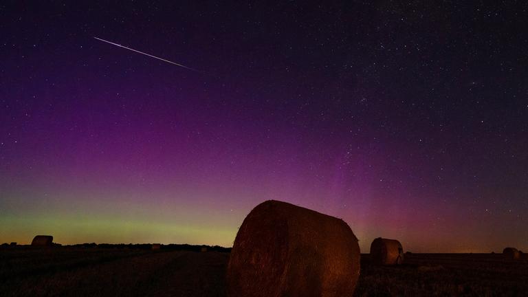 Hinter Strohballen leuchten Nordlichter, während eine Sternschnuppe am Himmel verglüht. Perseiden erreichen heute ihren Höhepunkt über dem Brandenburger Himmel