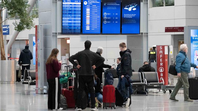 Passagiere stehen in der Flughafenhalle und informieren sich an einer Anzeigetafel.