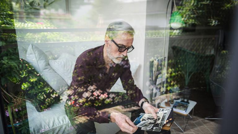 Blick durch eine spiegelnde Scheibe: Ein Mann mit geraumeliertem Haar, Bart und dunkler Brille sitzt auf einem Sofa und betrachtet Fotos.