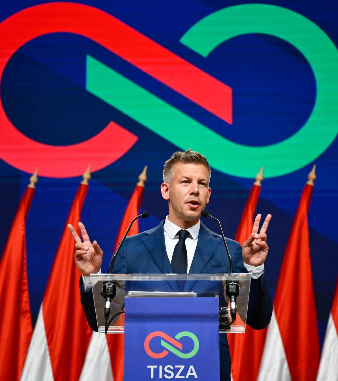 Peter Magyar auf einer Bühne. zeigt seine beiden Hände im Stile des Victory-Zeichens neben sich. Hinter ihm das Tisza-Logo.