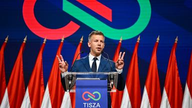 Peter Magyar auf einer Bühne. zeigt seine beiden Hände im Stile des Victory-Zeichens neben sich. Hinter ihm das Tisza-Logo.