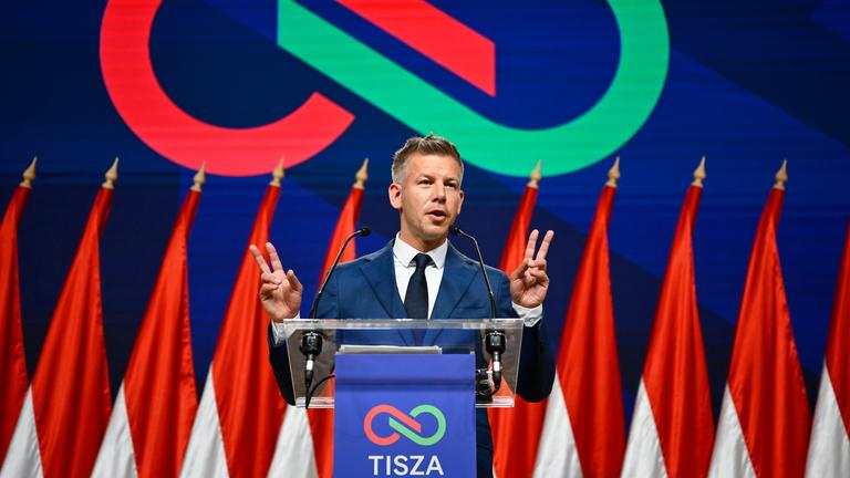 Peter Magyar auf einer Bühne. zeigt seine beiden Hände im Stile des Victory-Zeichens neben sich. Hinter ihm das Tisza-Logo.