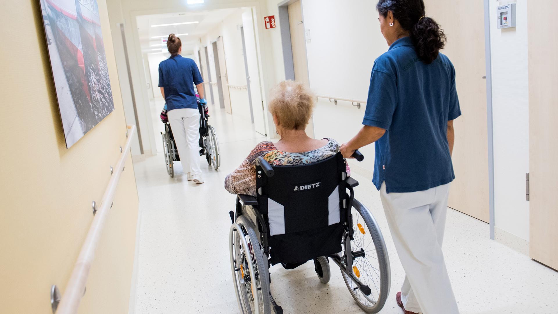 Zwei Pflegerinnen schieben in einem Klinikum Patienten mit Rollstühlen durch einen Flur.