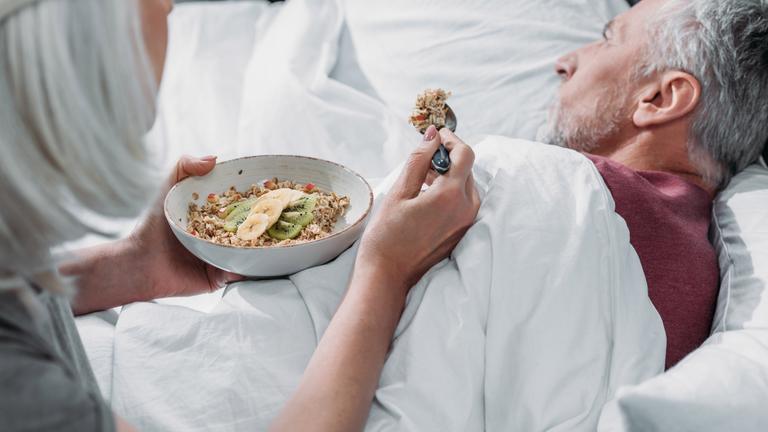 Ältere Frau mit grauen Haaren hält eine Schüssel mit Cerealien und Obst in der einen Hand und einen Löffel in der anderen, um damit ihren kranken Ehemann zu füttern, der von ihr weggedreht im Bett liegt