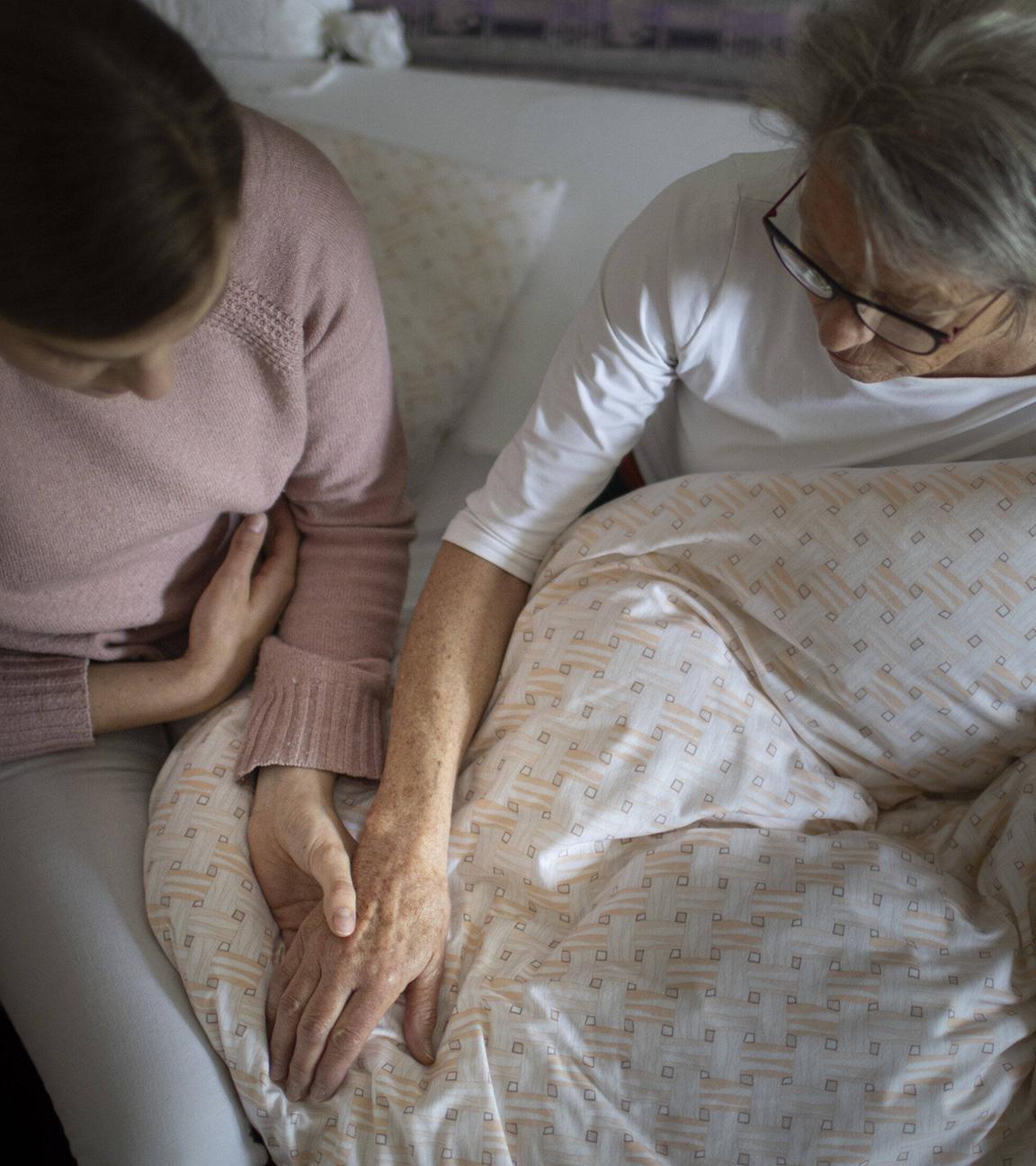 Eine junge Frau sitzt auf der Bettkante eines Bettes und hält die Hand einer Seniorin.