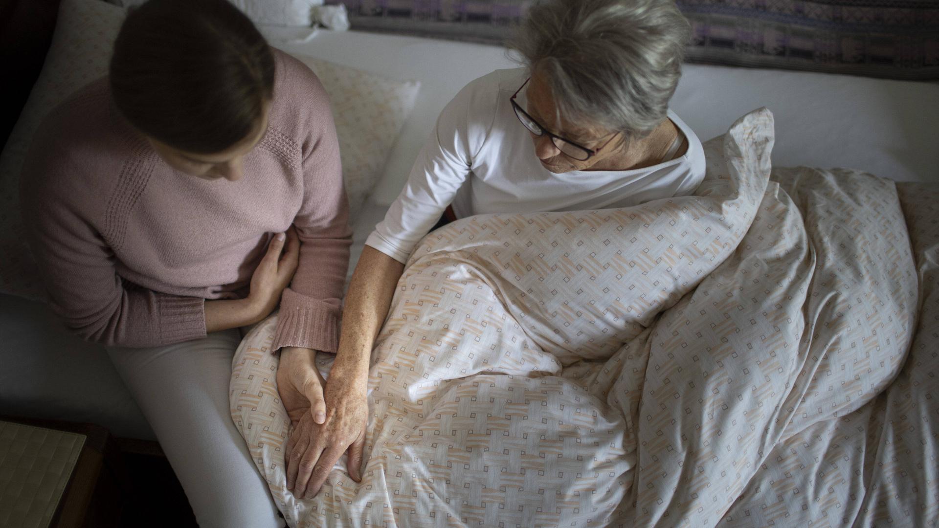 Eine junge Frau sitzt auf der Bettkante eines Bettes und hält die Hand einer Seniorin.