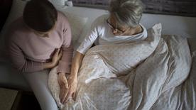 Eine junge Frau sitzt auf der Bettkante eines Bettes und hält die Hand einer Seniorin.
