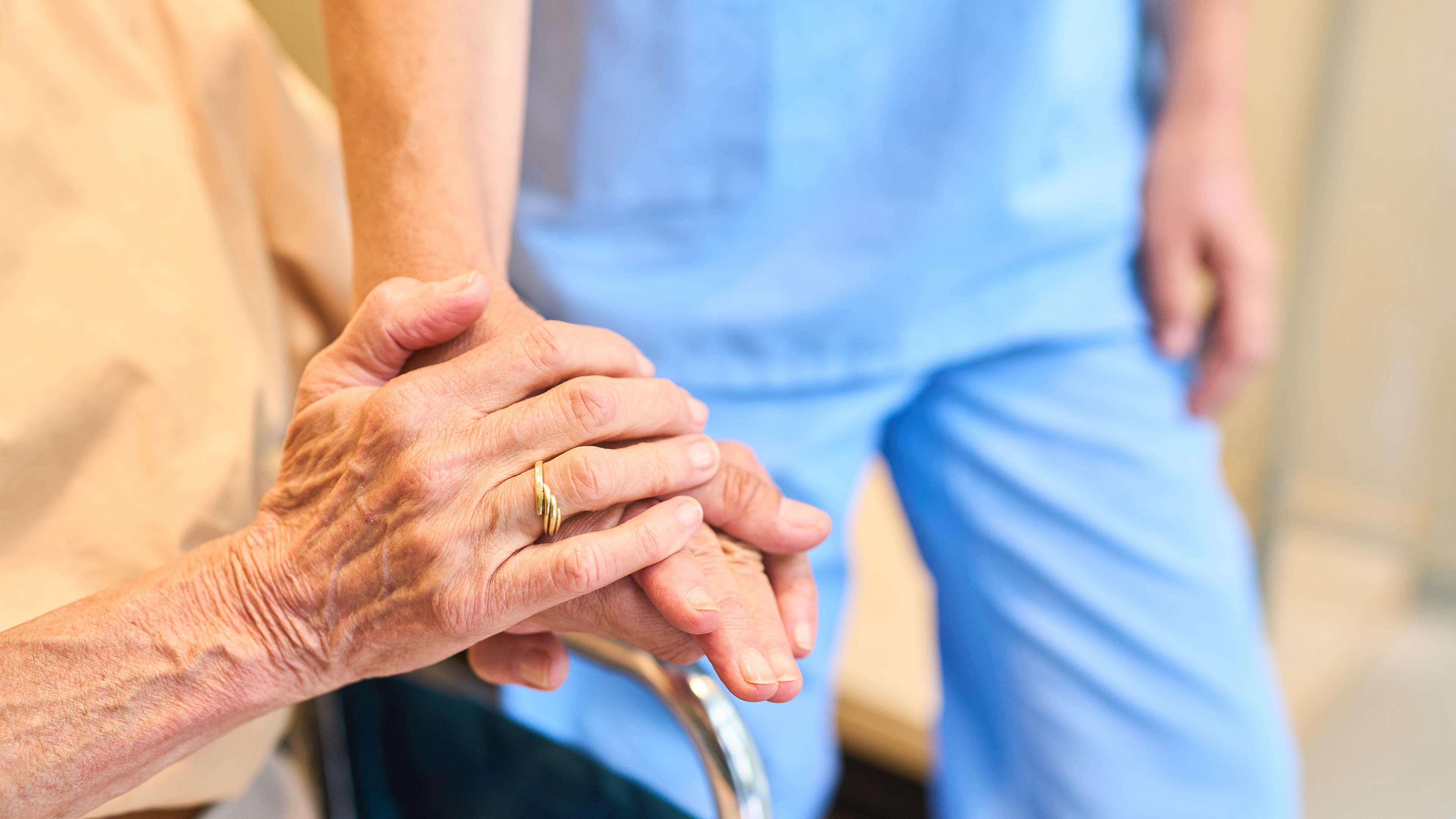 Ältere Person im Rollstuhl hält die Hand einer Pflegefachkraft.