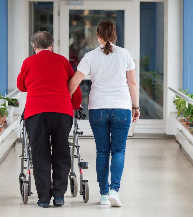 pflegerin fuehrt seniorin mit rollator durch einen flur