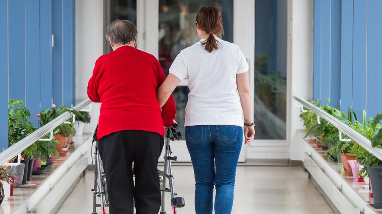 pflegerin fuehrt seniorin mit rollator durch einen flur