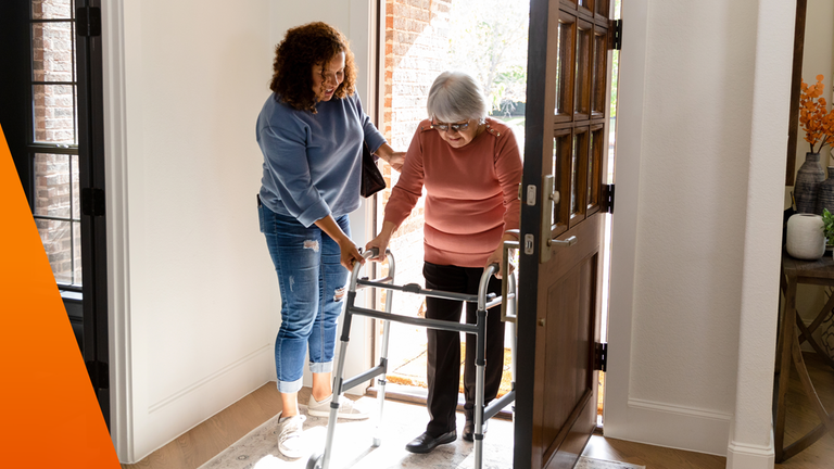 Ältere Frau schiebt Rollator in Eingangstür einer Wohnung, eine andere Frau hilft ihr dabei.