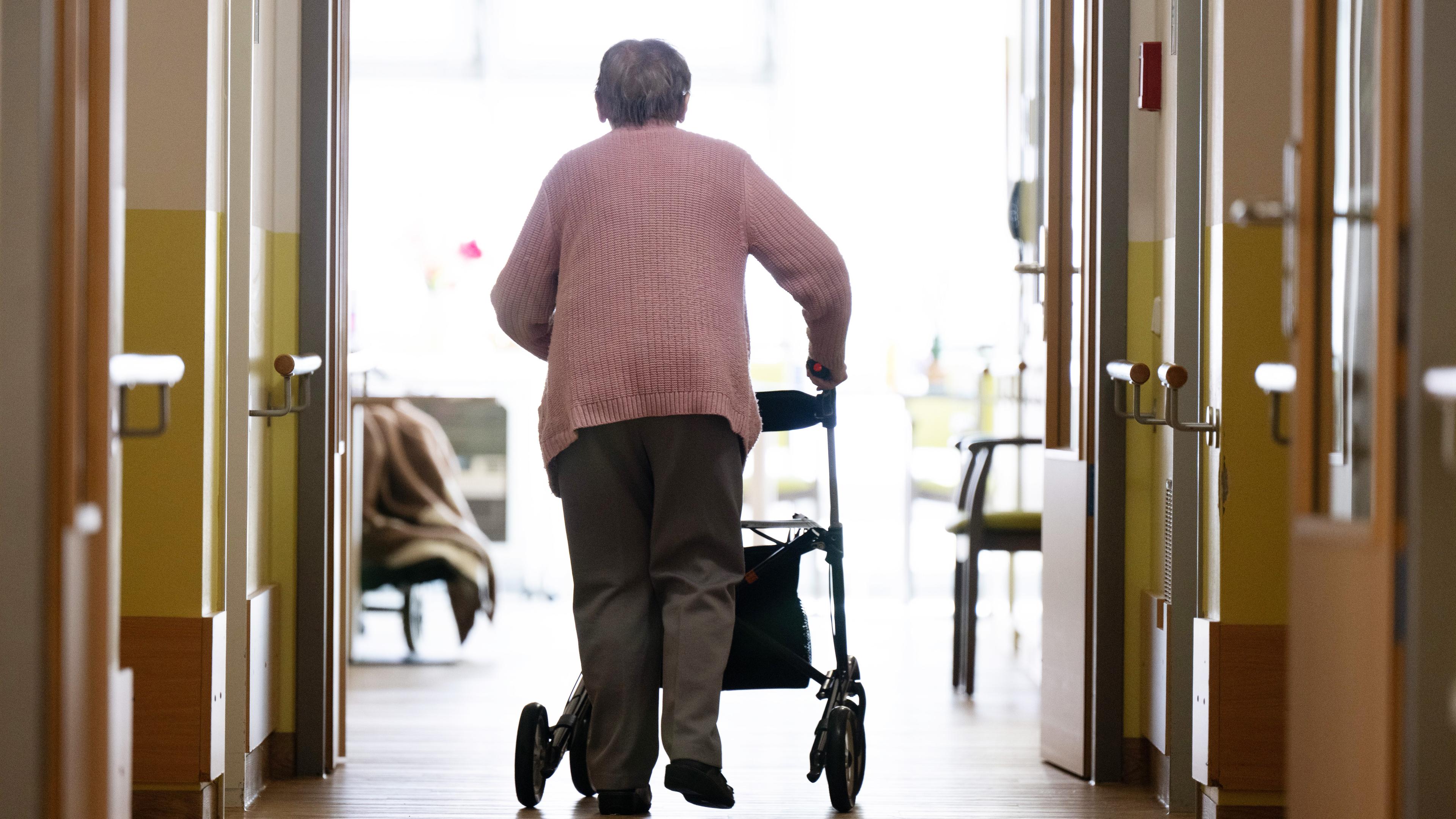 Eine Frau geht mit einem Rollator in einem Altenheim auf einem Gang entlang.
