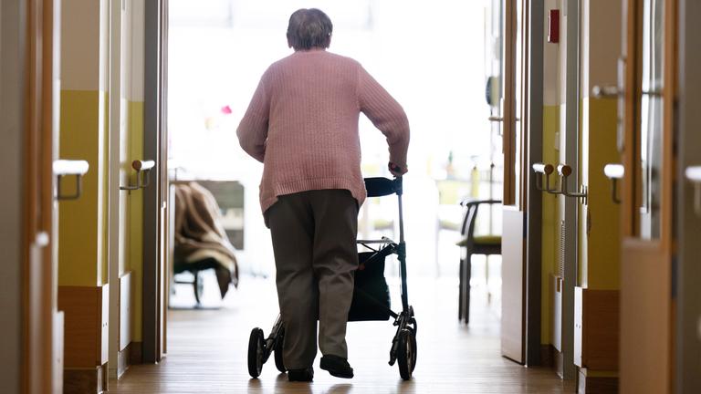 Eine Frau geht mit einem Rollator in einem Altenheim auf einem Gang entlang.