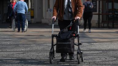 Eine ältere Person geht mit einem Rollator über einen gepflasterten Platz.