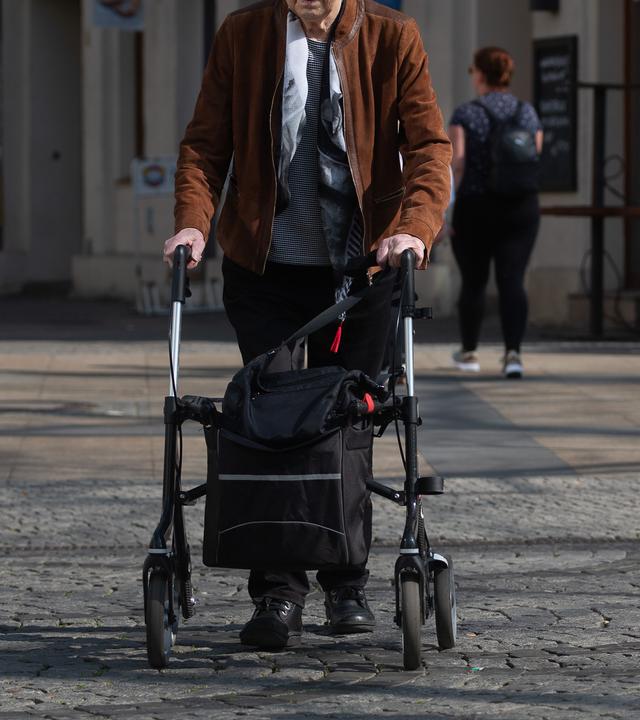 Eine ältere Person geht mit einem Rollator über einen gepflasterten Platz.