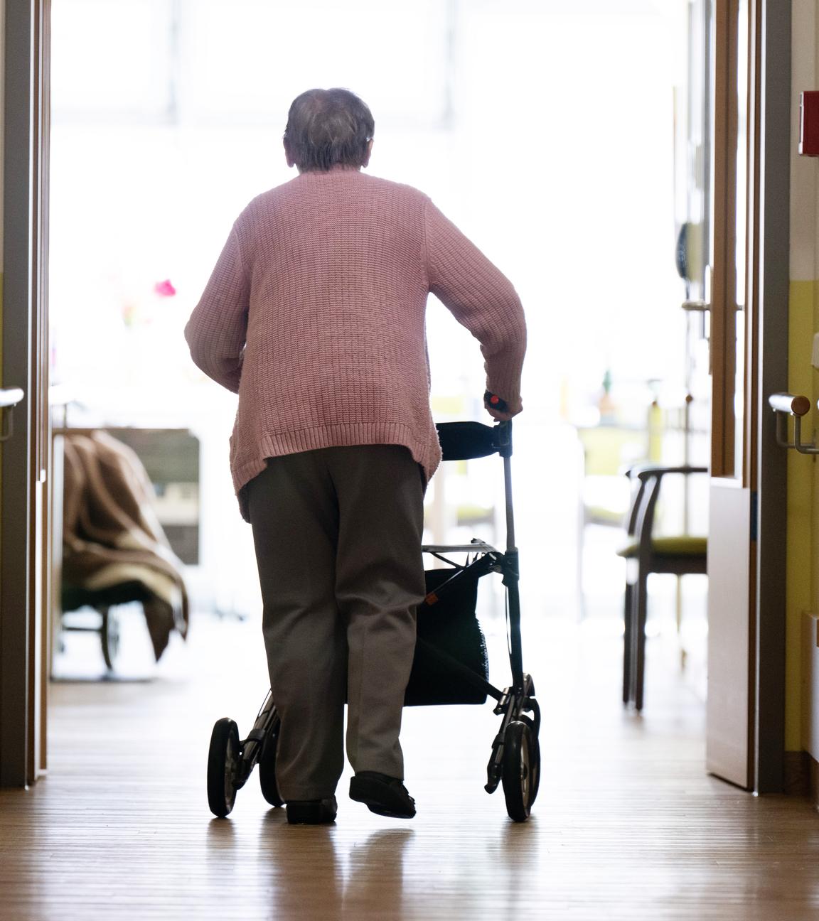 Eine Frau geht mit einem Rollator in einem Altenheim auf einem Gang entlang.