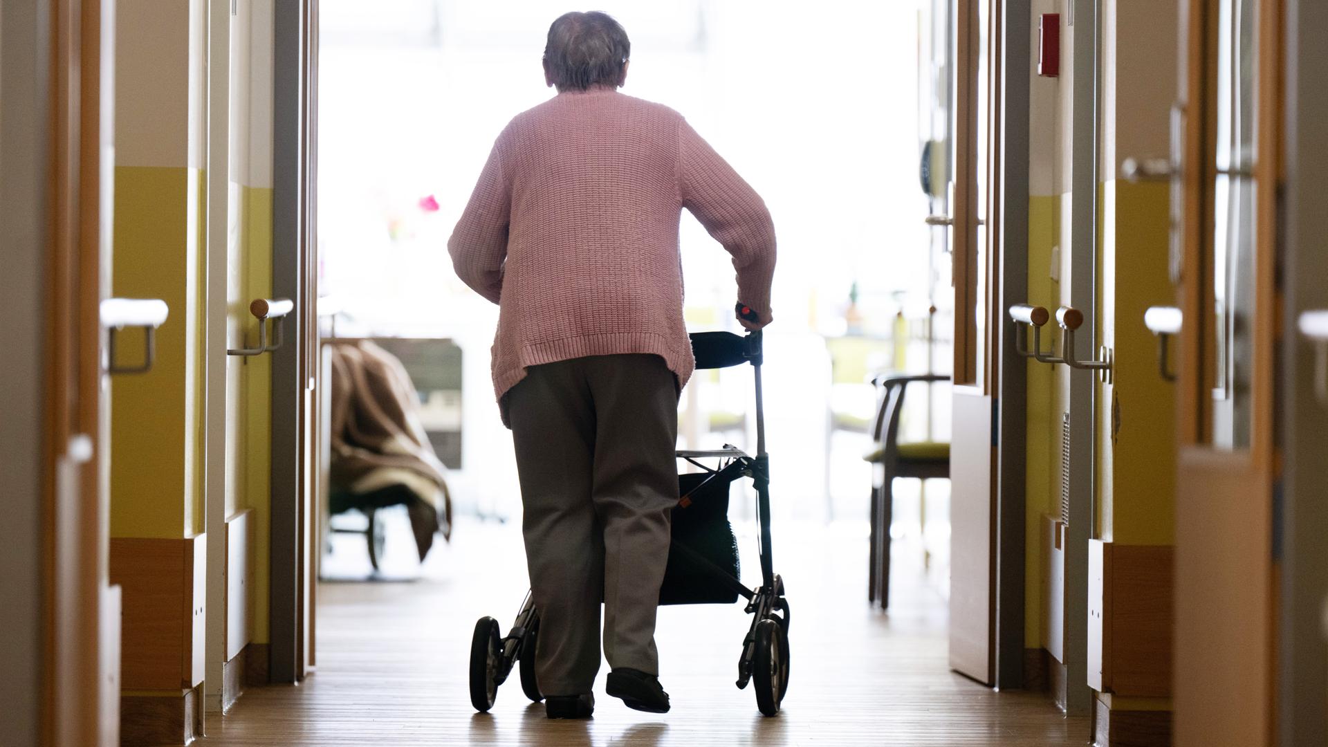 Eine Frau geht mit einem Rollator in einem Altenheim auf einem Gang entlang.