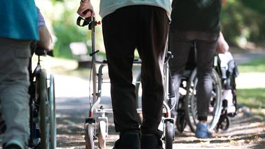 Mehrere Senioren sind mit Rollator oder Rollstuhl in einem Park spazieren.