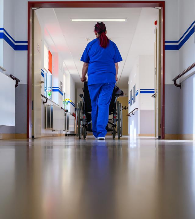 Eine Pflegerin der Geriatrie schiebt einen Patienten im Rollstuhl über die Station.
