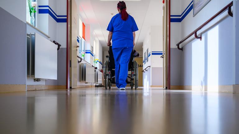 Eine Pflegerin der Geriatrie schiebt einen Patienten im Rollstuhl über die Station.