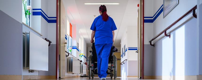 Eine Pflegerin der Geriatrie schiebt einen Patienten im Rollstuhl über die Station.