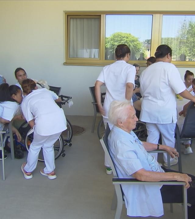 Personen sitzen in einem Pflegeheim an einem Tisch. Es laufen mehrere Ärzte herum und sitzen ebenfalls am Tisch.