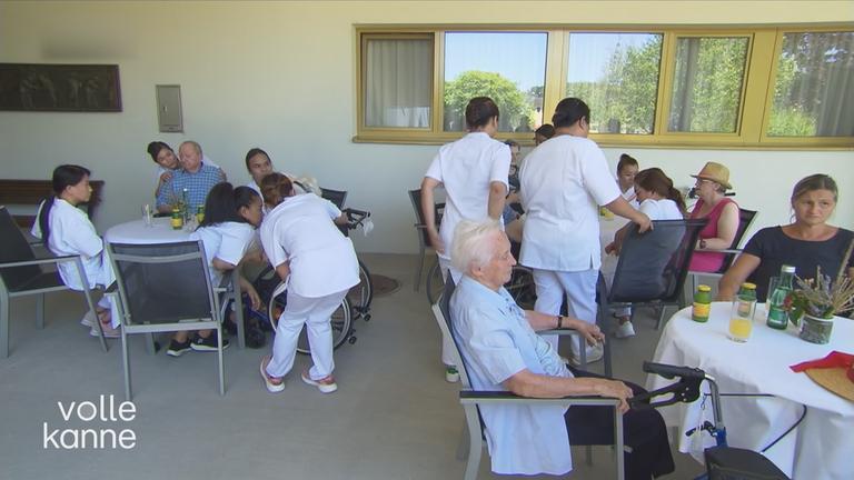 Personen sitzen in einem Pflegeheim an einem Tisch. Es laufen mehrere Ärzte herum und sitzen ebenfalls am Tisch.