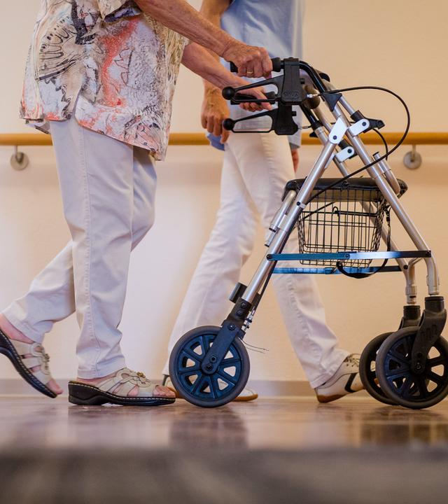 Eine alte Dame schiebt ihren Rollator und wird von einer Pflegekraft begleitet. 