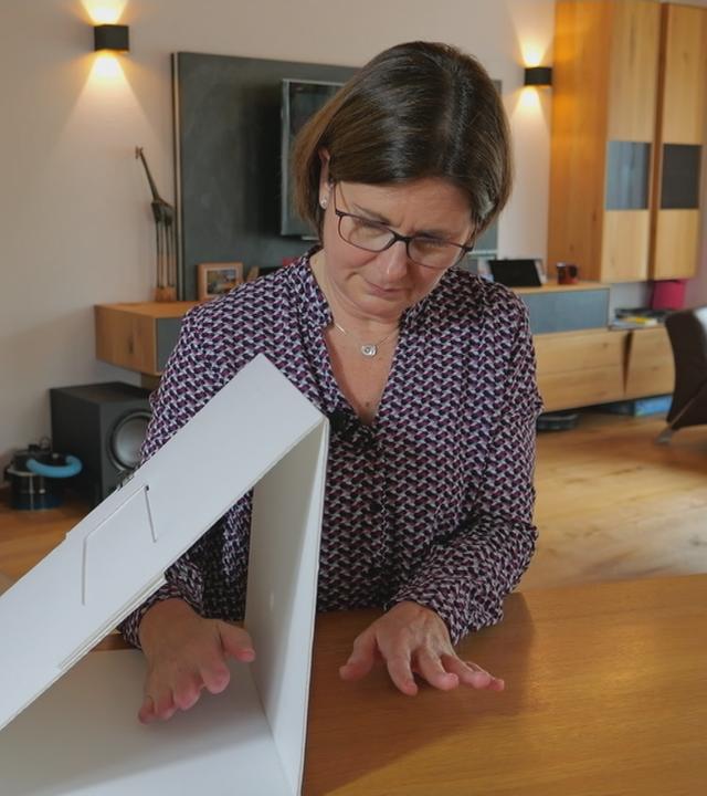 Eine Frau hält eine Hand neben eine Art Karton und die andere Hand (bei der die Extremitäten nicht mehr völlig vorhanden sind) in den Karton