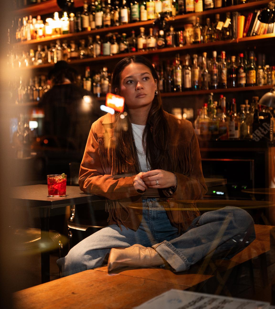 Leonie, eine junge Frau, sitzt in einer Bar auf einer Bank und schaut gedankenverloren aus dem Fenster. Sie stützt sich auf einem Tisch ab, auf dem ein Getränk steht.