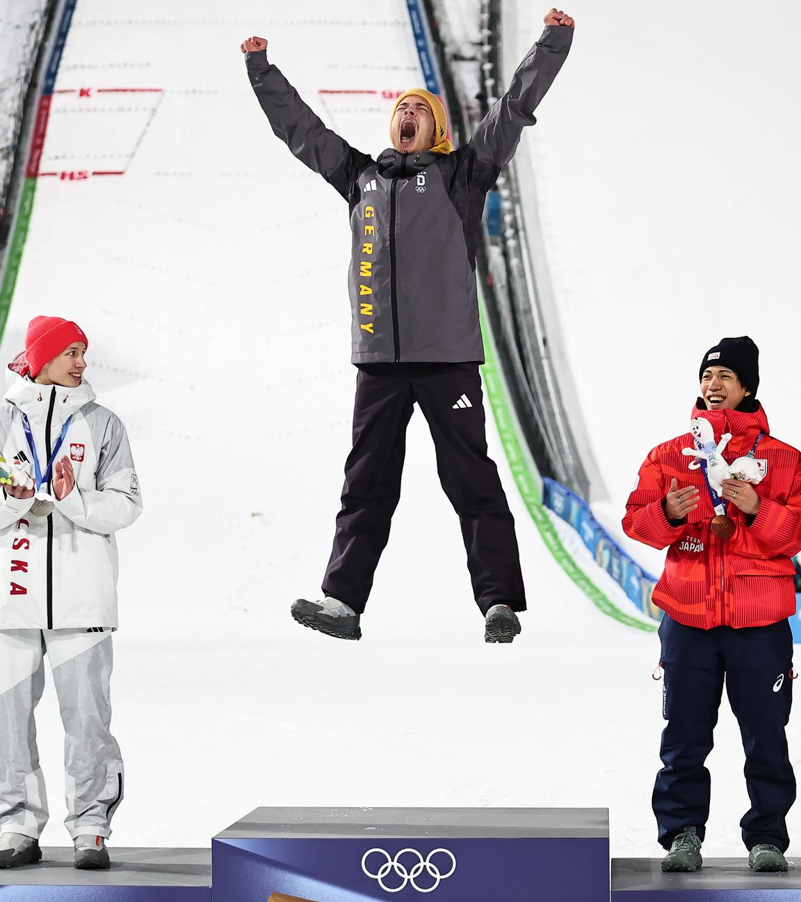 Olympiasieger Philipp Raimund (GER) jubelt bei der Siegerehrung auf der Normalschanze neben Kacper Tomasiak (POL), Ren Nikaido (JPN) und Gregor Deschwanden (SUI) über seine Goldmedaille.