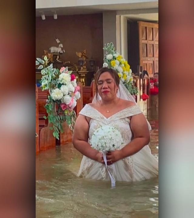 Ein Braut in weißem Kleid und Brautstrauß in der Hand läuft in einer überschwemmten Kirche zum Altar, das Wasser steht bis über ihre Knie.