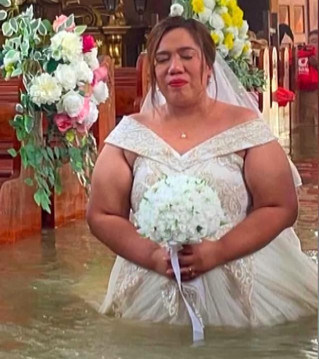 Ein Braut in weißem Kleid und Brautstrauß in der Hand läuft in einer überschwemmten Kirche zum Altar, das Wasser steht bis über ihre Knie.