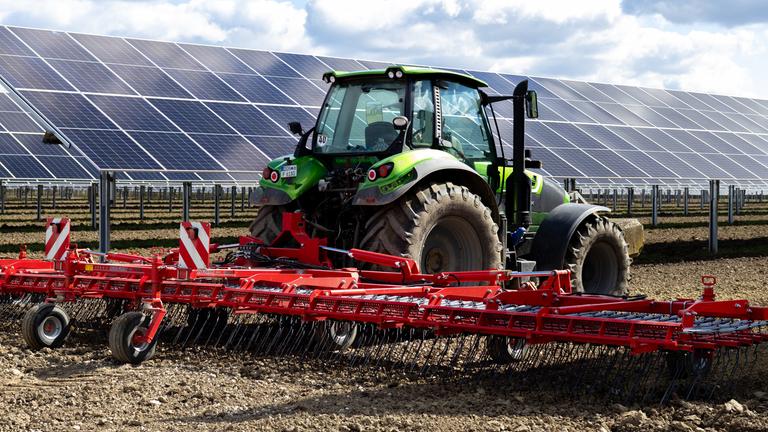 Ein Traktor wird auf einem Feld eingesetzt, dahinter stehen Solarzellen.
