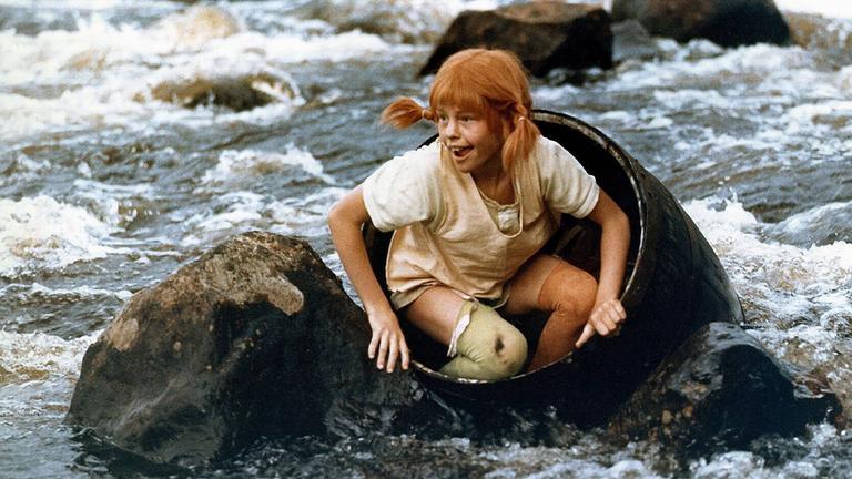 Pippi Langstrumpf sitzt in einem Fass, das in einem Fluss schwimmt. Es ragen Felsen aus dem Wasser. 