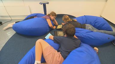 Schulkinder liegen auf blauen Sitzsäcken auf dem Boden und beschäftigen sich mit Tablets in der Schule