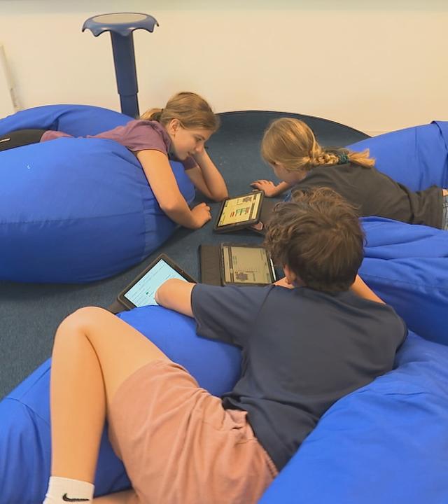 Schulkinder liegen auf blauen Sitzsäcken auf dem Boden und beschäftigen sich mit Tablets in der Schule