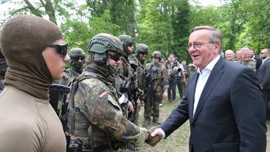 Verteidigungsminister Boris Pistorius schüttelt einer Bundeswehrsoldatin in schwerer Ausrüstung die Hand, beide lächeln. Am linken Bildrand steht ein Soldat mit Sonnenbrille und brauner Sturmhaube.