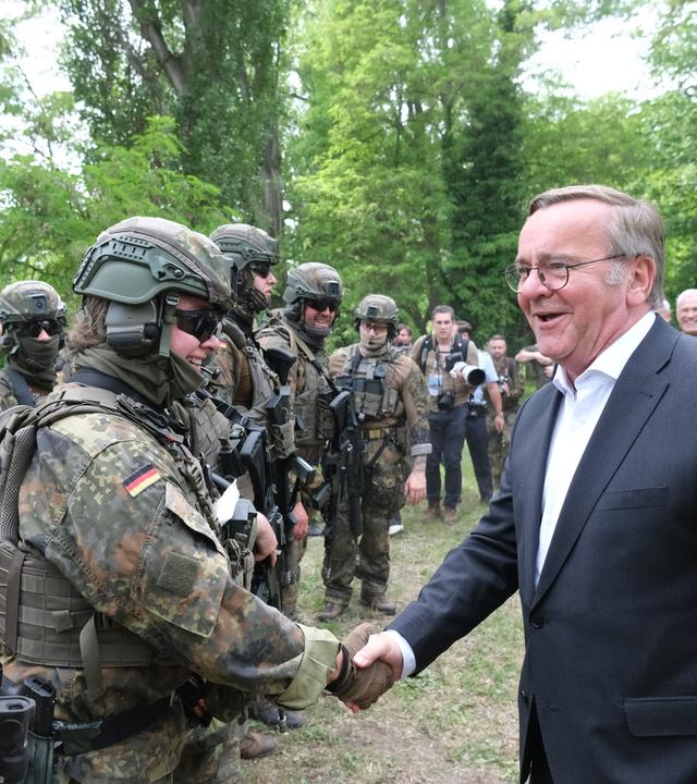 Verteidigungsminister Boris Pistorius schüttelt einer Bundeswehrsoldatin in schwerer Ausrüstung die Hand, beide lächeln. Am linken Bildrand steht ein Soldat mit Sonnenbrille und brauner Sturmhaube.